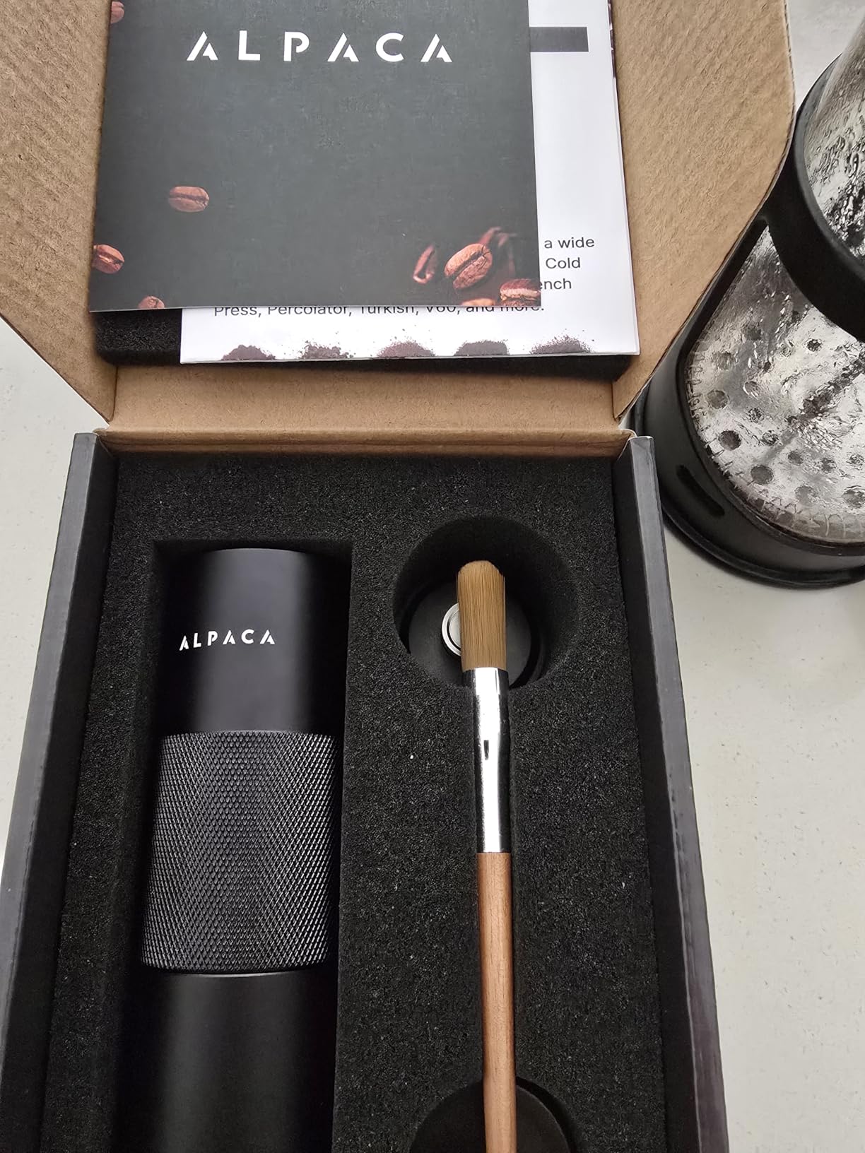 Opened Alpaca box with the black grinder and cleaning brush in a foam insert