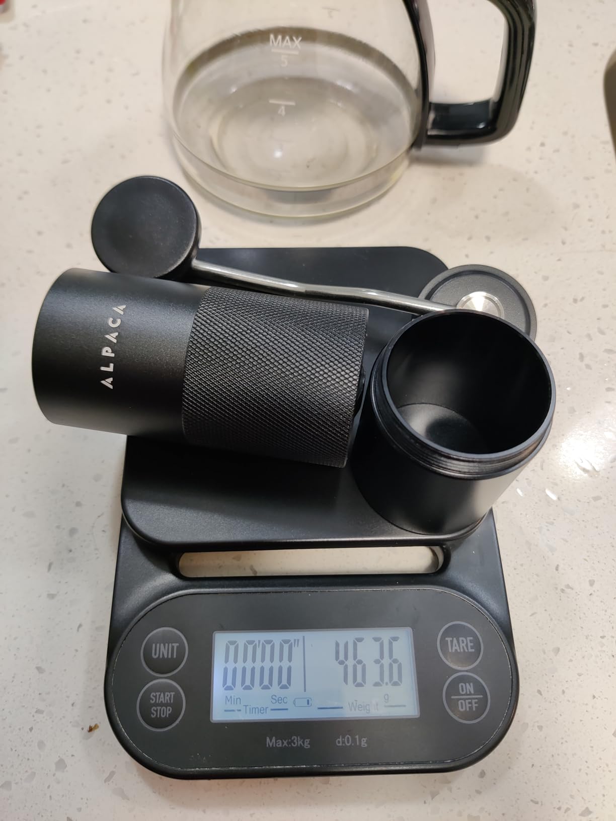 Black Alpaca grinder and its catch cup on a kitchen scale beside a glass coffee pot